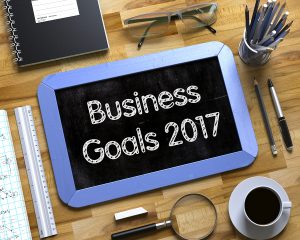 EPoS Technology - Business Goals 2017 on Small Chalkboard. Business Goals 2017 Handwritten on Blue Small Chalkboard. Top View of Wooden Office Desk with a Lot of Business and Office Supplies on It. 3d Rendering.
